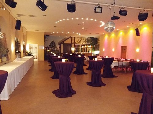 Der Saal des Gasthauses ist festlich hergerichtet und beleuchtet: In der Mittte stehen lila behusste Bartische mit Teelichtern, im Hintergrund stehen festlich eingedeckte Esstische mit Stühlen.