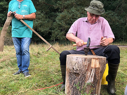 Ein älterer Mann sitzt vor einem Dengelbock und bearbeitet ein Sensenblatt mit dem Dengelhammer. (Foto © Inka Schwand)