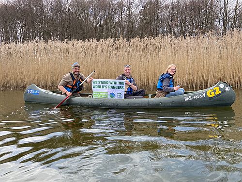 Drei Ranger sitzen während einer Kanutour im Boot, einer von ihnen hält ein kleines Banner hoch anlässlich des Welt-Ranger-Tags