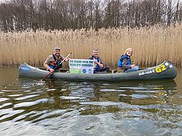 Drei Ranger sitzen während einer Kanutour im Boot, einer von ihnen hält ein kleines Banner hoch anlässlich des Welt-Ranger-Tags