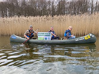 Drei Ranger sitzen während einer Kanutour im Boot, einer von ihnen hält ein kleines Banner hoch anlässlich des Welt-Ranger-Tags